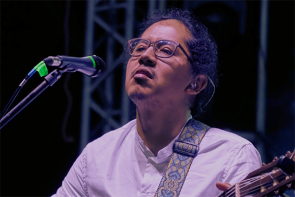 A headshot photo of Ati Cachimuel wearing a white shirt and glasses and singing into a microphone while playing the guitar. 