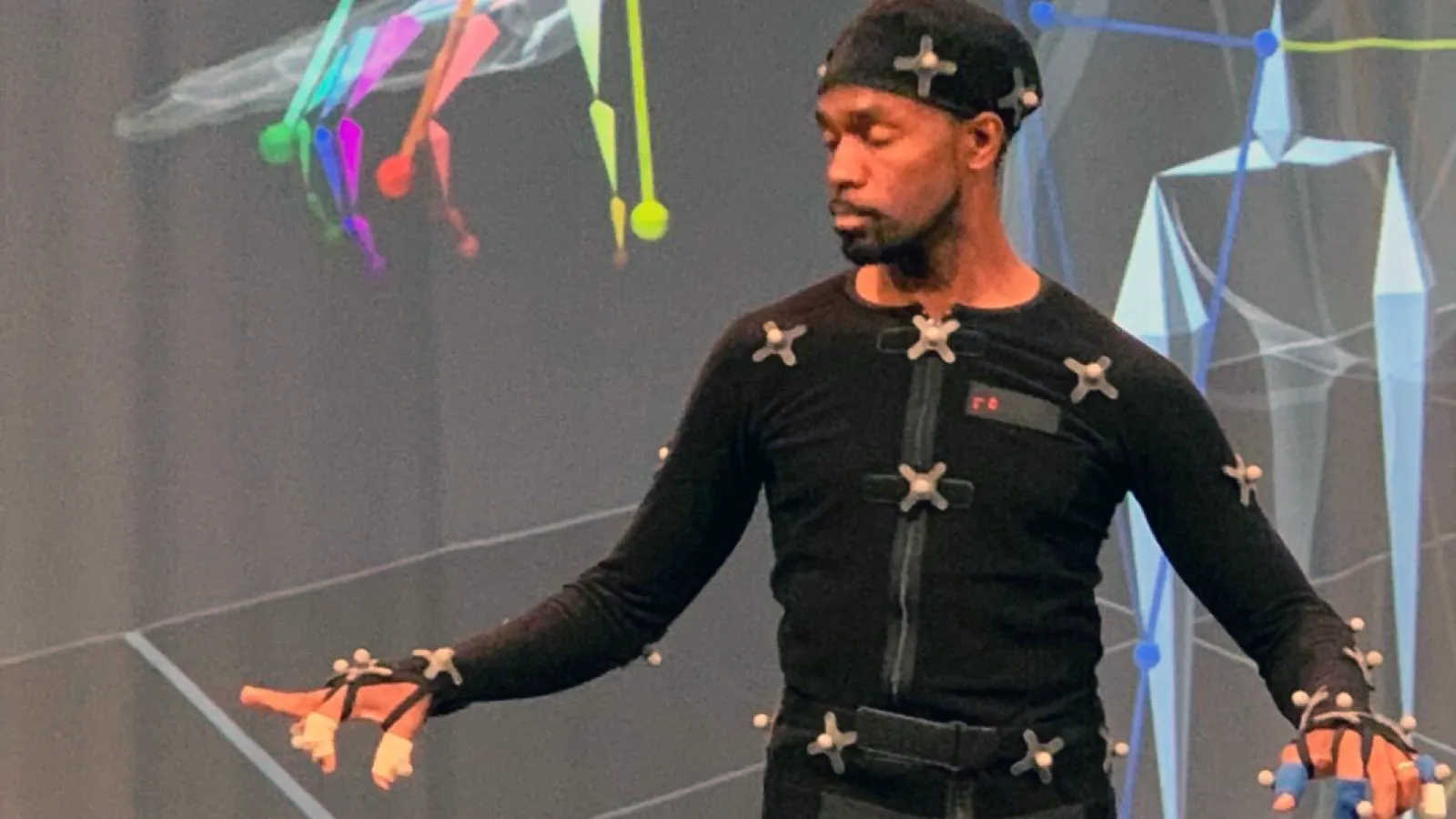 André Zachery suited up in the ACCAD Motion Lab. He looks down at the motion trackers on his fingers while his avatar is partially in view in the background.
