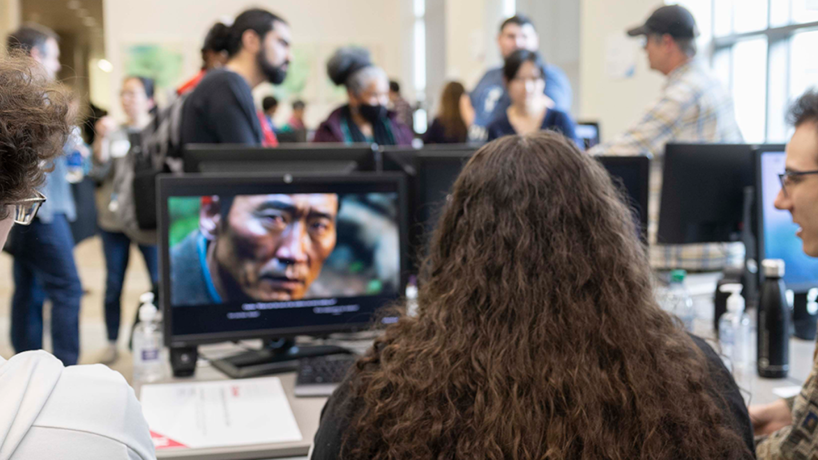 A group of three students sit at a table, watching a student-made video on a desktop computer. In the background, people socialize in the ACCAD computer lab during the Open House.