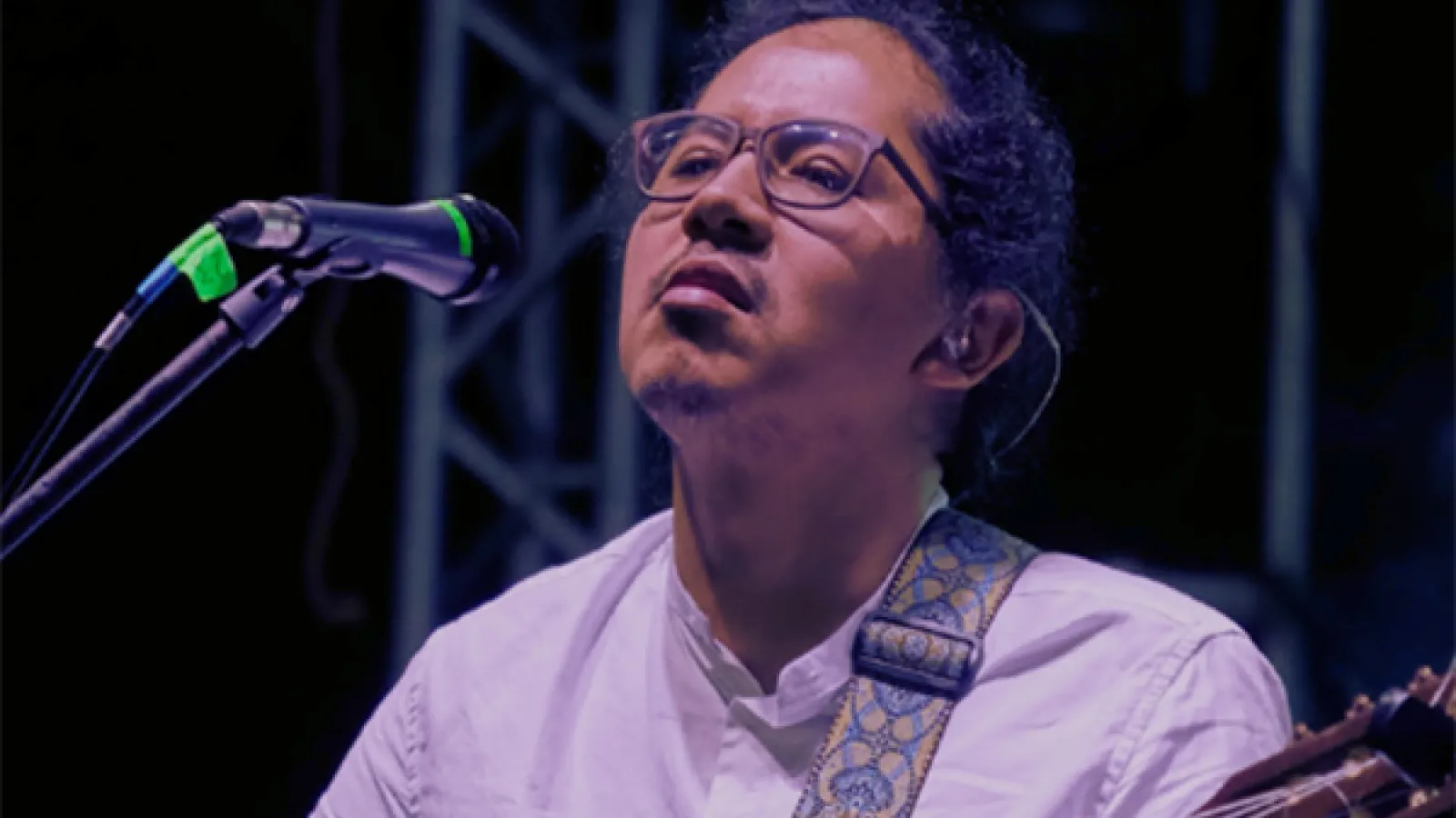 A headshot photo of Ati Cachimuel wearing a white shirt and glasses and singing into a microphone while playing the guitar. 