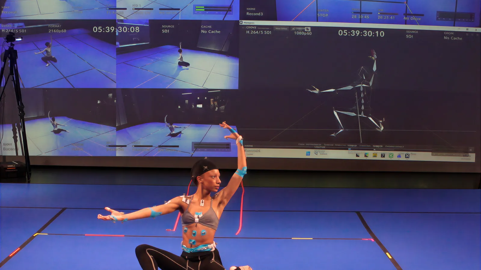A dancer wearing motion capture markers poses on a blue studio floor. Behind them, a large display wall shows multiple live video feeds and a real-time 3D skeletal wireframe tracking their movement.