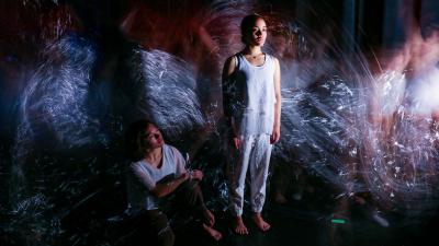 Two women, one sitting and one standing, pause in stillness as choatic light blur swirl around them